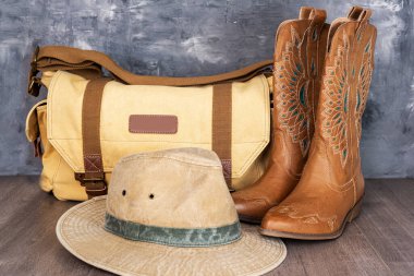 Leather cowgirl boots with a pattern, a hat and a canvas bag are on the floor against a gray wall.