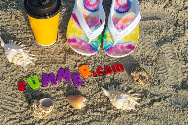 beach slippers, a cup of coffee and sea shells on the beach sand with the inscription SUMMER. In the rays of the morning sun.