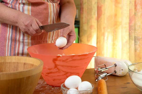 hostess in an apron breaks a raw egg into a bowl of flour with a knife to make dough. ingredients for cooking on the countertop