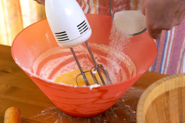 hostess in the home kitchen with a mixer pours sugar into a bowl for making a cake. Close-up of hands.