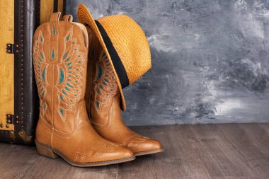 Leather cowgirl boots with straw hat pattern and an old suitcase stand on the floor against gray wall. Space for text.