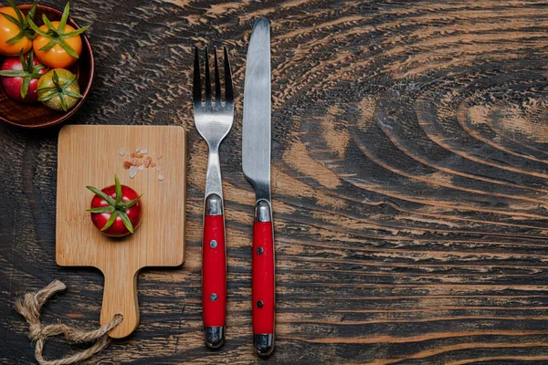 Stock image one ripe cherry tomatoes on cutting board.Fork and knife on dark wooden table. view from above. Copy space.