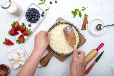 Aşçı süzme peyniri güveçte pişirme kabına koyar. Un, yumurta, merdane, tereyağı, süt ve çilek beyaz bir masada. Yukarıdan görüntüle.
