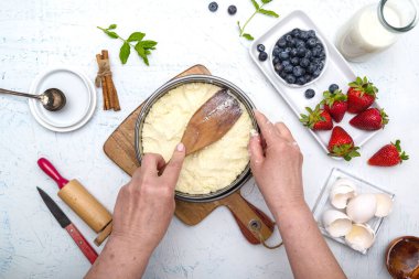 Aşçı süzme peyniri güveçte pişirme kabına koyar. Un, yumurta, merdane, tereyağı, süt ve çilek beyaz bir masada. Yukarıdan görüntüle.