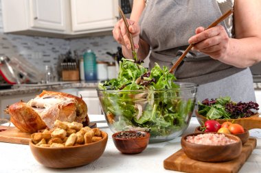 Kadın elleri ev mutfağında sebzeli ve tavuklu sezar salatası yapar. Önplanda kızarmış ekmek, tavuk ve tuz var..