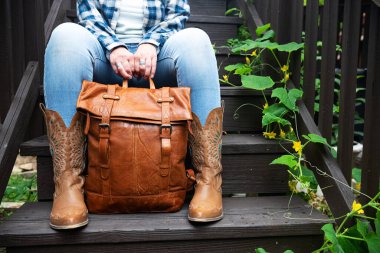 Kadın merdivenlerde otururken kahverengi deri bir sırt çantası tutuyor. Yakın plan eller ve deri kovboy çizmeleri..
