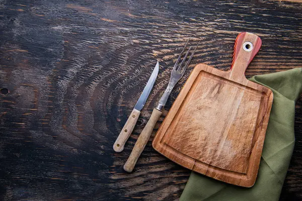 Wood cutting board with linen napkin, knife and fork on wooden table with copy space, top view