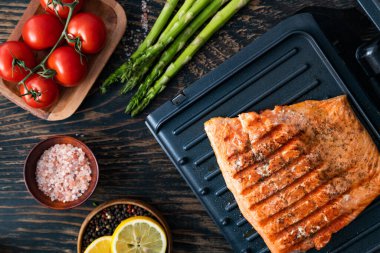 Tuz, biber ve otlarla tatlandırılmış elektrikli ızgarada somon filetosu. Domates, limon ve kuşkonmaz. Yukarıdan görüntüle.