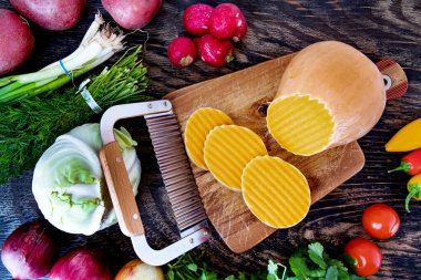 Kesme tahtasında dalga bıçağıyla kesilmiş bir balkabağı. Tahta bir masada çeşitli sebzeler. Yukarıdan görüntüle.