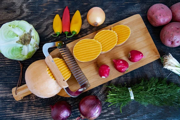 Kesme tahtasında dalga bıçağıyla kesilmiş bir balkabağı. Tahta bir masada çeşitli sebzeler. Yukarıdan görüntüle.
