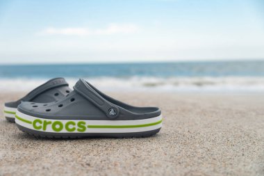 Gray Crocs sandals lie on the sand by the ocean. trendy croc beach shoes. Travel vacation banner background. clipart