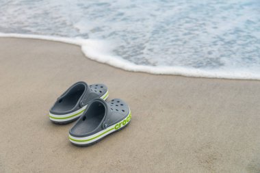 Gri Crocs sandaletleri okyanusun kıyısındaki kumda yatıyor. Moda timsah plajı ayakkabıları. Seyahat tatil afişi arkaplanı.