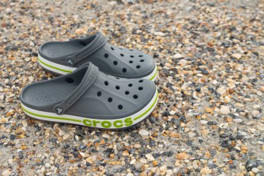 Gri Crocs sandaletleri kumlu sahilde yavaşça dinleniyor, pürüzsüz hatları dingin arkaplanı tamamlıyor.