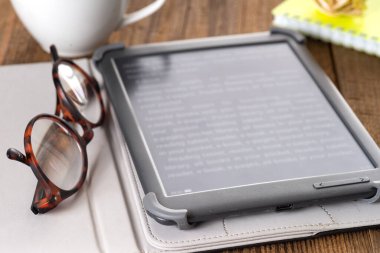 E-kitap, ahşap masanın üzerinde bir fincan kahve ve okuma gözlüğü.