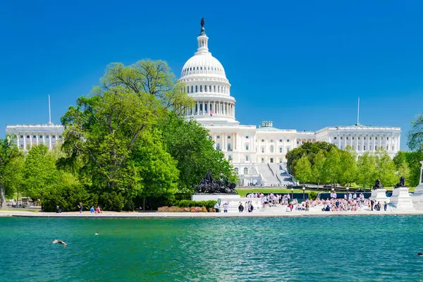 ABD Kongre Binası, yansıtma havuzunun önünden batıdan görüldü. Capitol yeşil ağaçlar ve mavi gökyüzü ile çevrilidir..