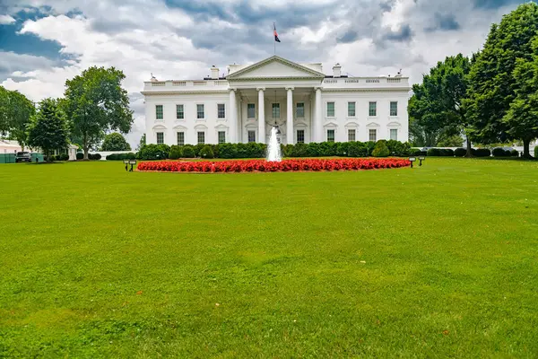 Bulutlu bir gökyüzünün altındaki Beyaz Saray. Kırmızı çiçekli ve çeşmeli büyük çimler..