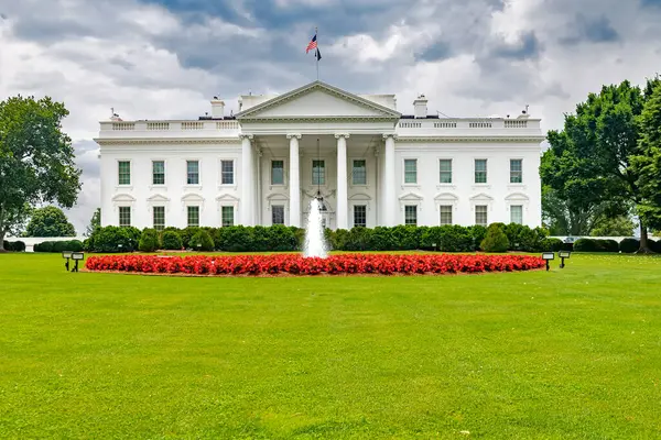 Bulutlu bir gökyüzünün altındaki Beyaz Saray. Kırmızı çiçekli ve çeşmeli büyük çimler..