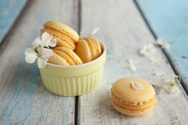 Ahşap mavi arka planda pastel sarı bir kasede Fransız kurabiyeli badem kurabiyesi. Fotokopi yeri olan pasta dükkanı kartı. Beyaz çiçekli geleneksel Fransız kurabiyeleri, bahar çiçekleri.