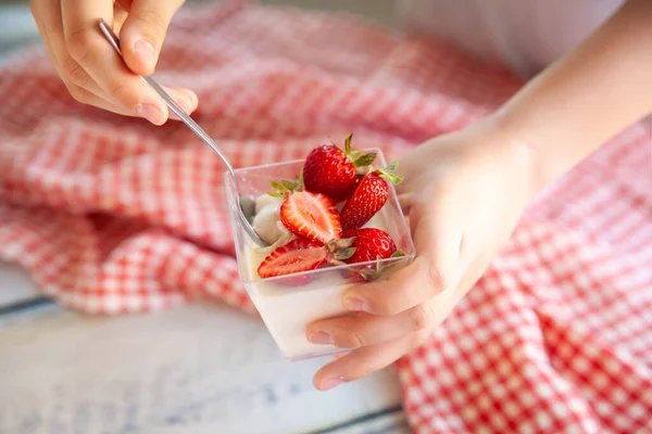 Çilek soslu vanilyalı panna cotta ve cam bardakta çilek. Ev yapımı tatlıyı tutan eller. Kırmızı ve beyaz kareli peçeteli açık tahta arkaplan