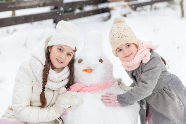 姐姐们在冬日的花园里和一个披着粉色针织围巾的雪人玩耍 — 图库照片