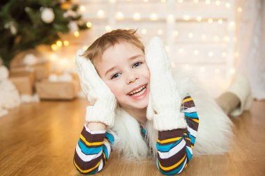 Noel arifesi. Sıcak beyaz kürk yelek giyen neşeli çocuk Noel süslemelerinde oynayan eldiven örüyor. Beyaz Noel konsepti