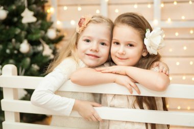 Noel arifesi. Tatil süsleriyle beyaz ahşap bankta oturan neşeli kızlar. Beyaz Noel konsepti