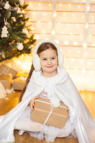A girl in white cape sitting among the winter decorations with Christmas tree and white toys. White Christmas concept