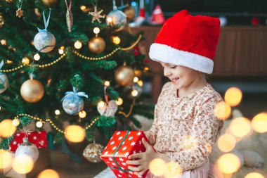 Kırmızı Noel Baba şapkalı şirin küçük kız elinde hediye kutusuyla Noel ağacının yanında oturuyor. Mutlu Noeller ve mutlu yıllar konsepti.