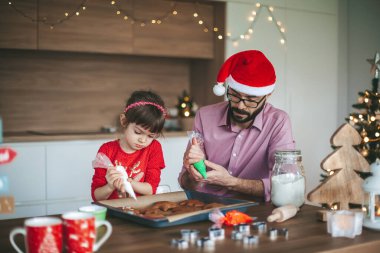 Yoğunlaşmış küçük kız ve babası zencefilli Noel kurabiyesi süslüyor. Aile Noel kutlaması için hazırlanıyor..