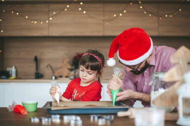 Sevimli küçük kız ve babası mutfakta zencefilli kurabiye süslüyor. Aile Noel ve Yeni Yıl için hazırlanıyor.