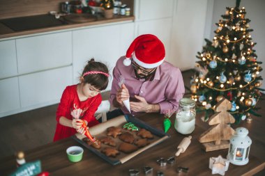 Noel Baba şapkalı baba ve küçük kızı zencefilli Noel kurabiyesi süslüyor.