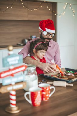Gülümseyen küçük kız ve babası Noel Baba 'nın şapkasıyla zencefilli Noel kurabiyesi süslüyor. Noel ve Yeni Yıl hazırlıkları.