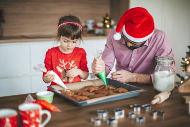 Küçük kız ve babası zencefilli Noel kurabiyesi dekore ediyorlar. Aile Noel ve Yeni Yıl kutlamaları için hazırlanıyor.