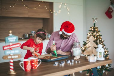 Sevimli küçük kız ve Noel Baba şapkalı babası zencefilli Noel kurabiyesi süslüyorlar.