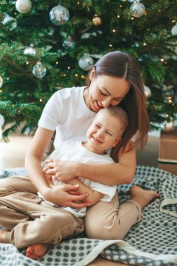 Güzel, gülümseyen bir kadın evdeki süslü Noel ağacının yanında battaniyenin üzerinde mutlu oğluna sarılıyor. Aileyle yeni yıl tatili.