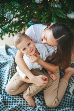 Güzel, gülümseyen küçük bir çocuk ve annesi süslü Noel ağacının altında birbirlerine sarılıyorlar. Aileyle yeni yıl tatili.