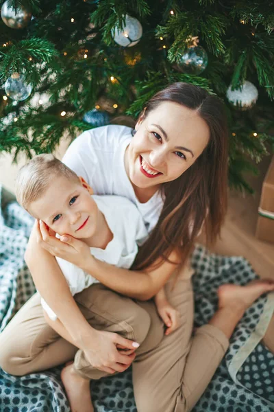 Güzel, gülümseyen bir kadın ve küçük oğlu evdeki süslü Noel ağacının altında sarılıyorlar. Aileyle yeni yıl tatili.