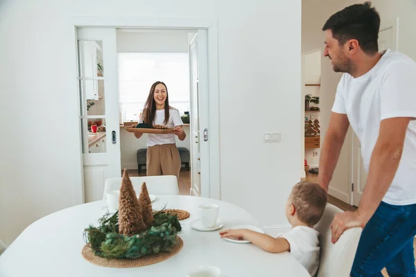 在圣诞节的早上 一个带着托盘和早餐的年轻女人在微笑 她的小儿子和她的丈夫坐在桌旁等她 — 图库照片