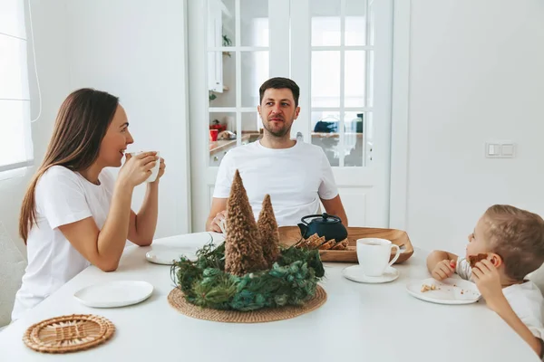 圣诞节早上 身穿白色T恤衫的一家人坐在桌旁 爸爸和他们的小儿子喝茶 吃蛋糕当圣诞早餐 — 图库照片