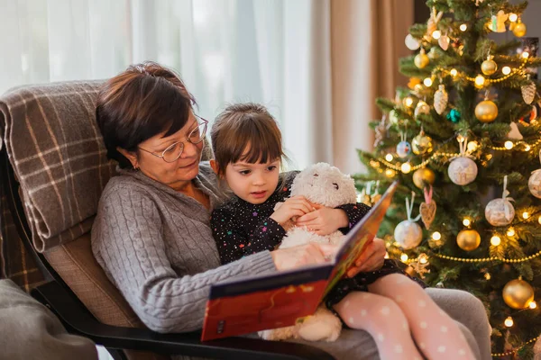 Küçük Kız Büyükannesi Noel Ağacının Yanındaki Sandalyede Otururken Birlikte Kitap — Stok fotoğraf