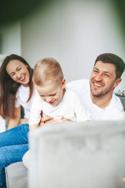 Küçük oğluyla mutlu, genç bir aile, yumuşak, gri bir kanepede oturuyor.