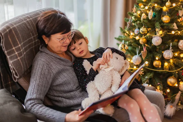 老妇人坐在圣诞树旁的椅子上给小孙女读书 — 图库照片