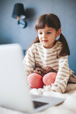 Cute little girl sitting on a bed at home and watching a video on a laptop
