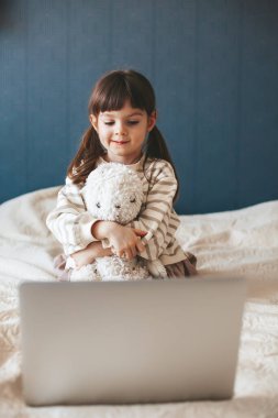 Little girl hugging her toy bunny sitting on a bed and watching something interesting on a laptop