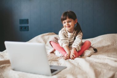 Yatakta oturmuş dizüstü bilgisayardan bir şeyler izleyen sevimli, gülümseyen küçük bir kız.