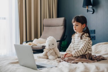 Serious adorable little girl sitting on a bed in a bedroom and watching something on a laptop