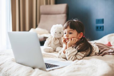 Cute little girl lying on a bed in a bedroom and watching something on a laptop