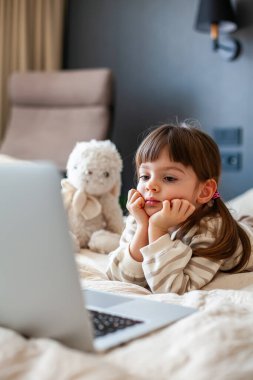 Portrait of a cute little girl lying on a bed in a bedroom and watching something on a laptop