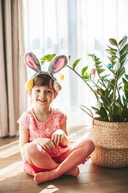 Cute little girl sitting near the plant decorated with Easter eggs. Happy playful child with bunny or rabbit ears. Easter concept.