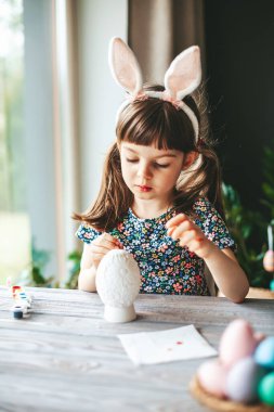Cute little girl with bunny ears sitting at the table painting Easter egg, made of gypsum